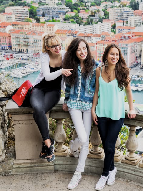 Three Sprachcaffe students smiling on a balcony overlooking Nice, France, with the harbour and colourful buildings around