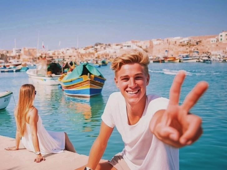 Girl sitting by the harbour in Malta, overlooking colourful traditional boats in the clear blue water.