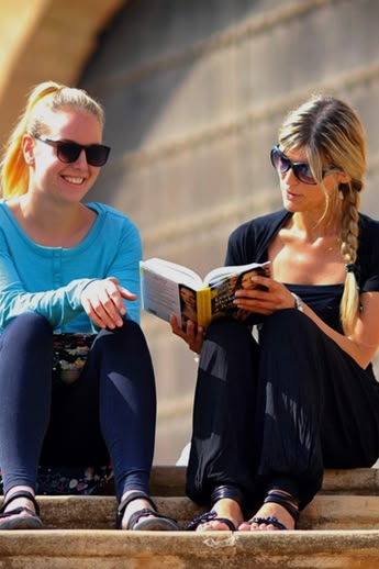 Two women sitting on stone steps reading a book in Rabat, Morocco, with historic architecture behind them.