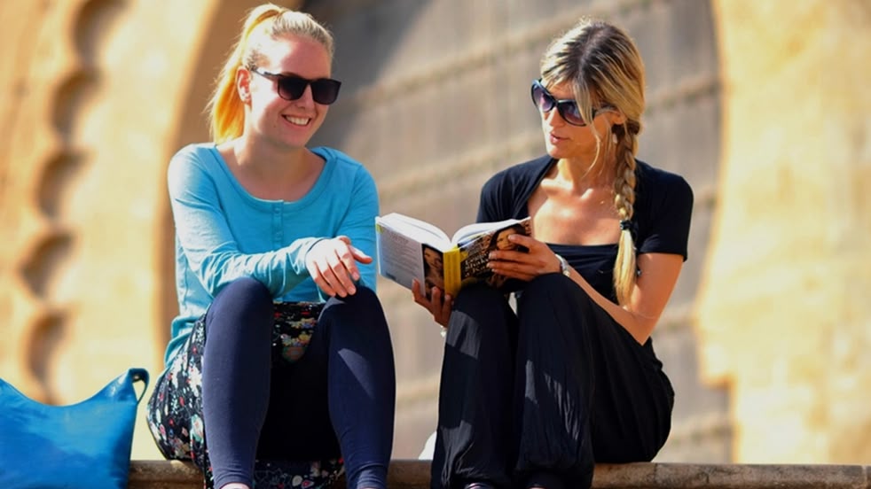 Two women sitting on stone steps reading a book in Rabat, Morocco, with historic architecture behind them.