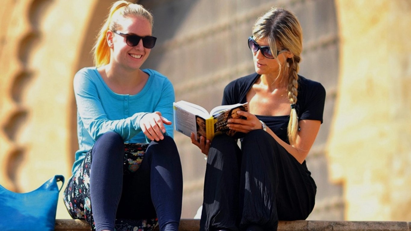 Two Sprachcaffe students sitting on steps in Rabat, Morocco, reading and chatting in front of traditional architecture.