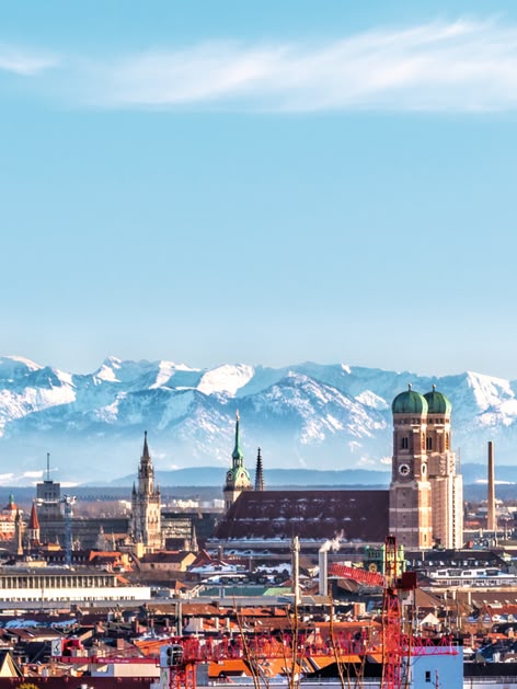 **ALT:** Panoramiczny widok na Monachium z Alpami w tle, ukazujący wieże Frauenkirche i inne zabytki miasta pod błękitnym niebem.