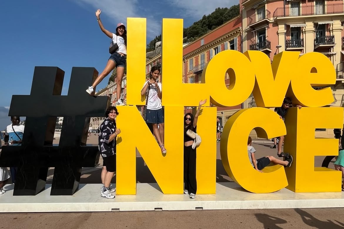Group of young people posing on and around the large yellow "#ILoveNICE" sign in Nice, France, on a sunny day.