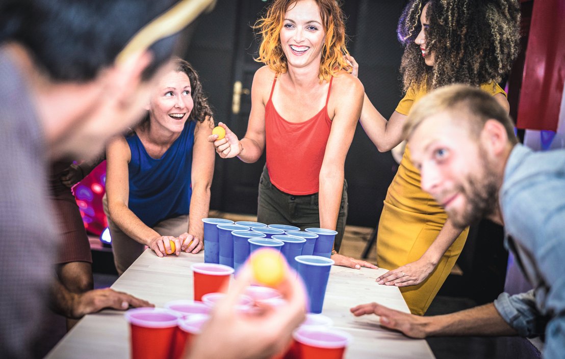 Hier haben Menschen Spaß beim Spielen während einer Party. Sie stehen vor einem Tisch. Im Fodergrund befindet sich ein orangener Ball zwischen den Fingern eines Menschen. Auf dem Tisch stehen rote und blaue Becher.