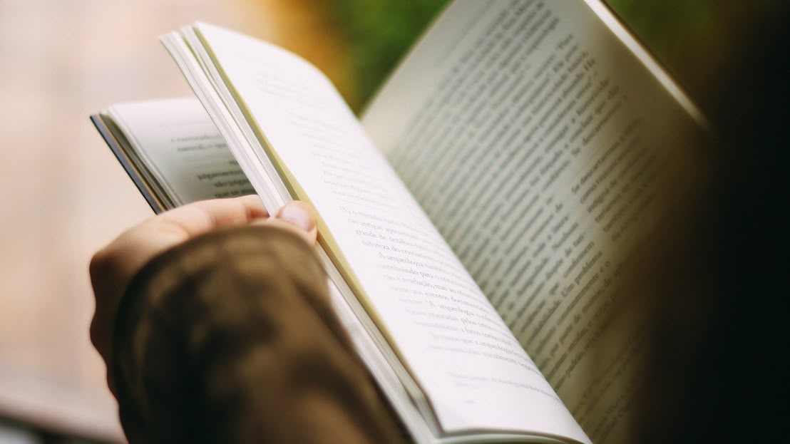 Людина тримає відкриту книгу, читаючи її. Видно лише одну руку з книгою, на фоні розмитого пейзажу.