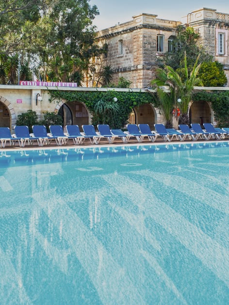 Large outdoor swimming pool surrounded by sun loungers and Mediterranean-style buildings at Sprachcaffe Malta.