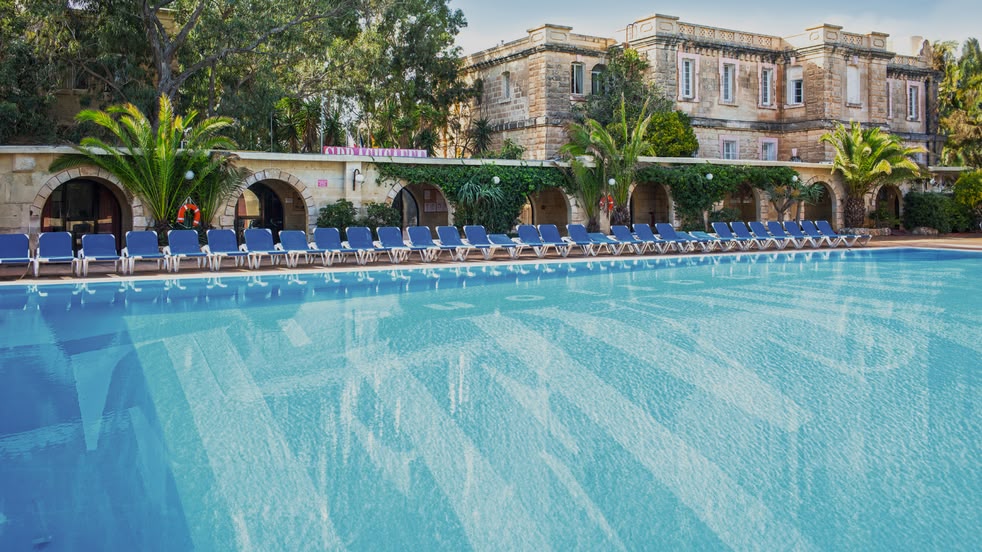 Large outdoor swimming pool surrounded by sun loungers and Mediterranean-style buildings at Sprachcaffe Malta.