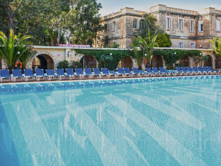 Large outdoor swimming pool surrounded by sun loungers and Mediterranean-style buildings at Sprachcaffe Malta.