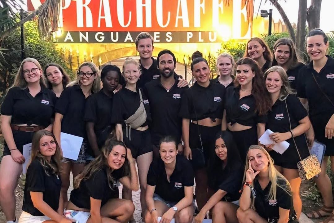 Grupa studentów Sprachcaffe pozuje przed szyldem szkoły, ubrani w czarne koszulki, w otoczeniu tropikalnych roślin