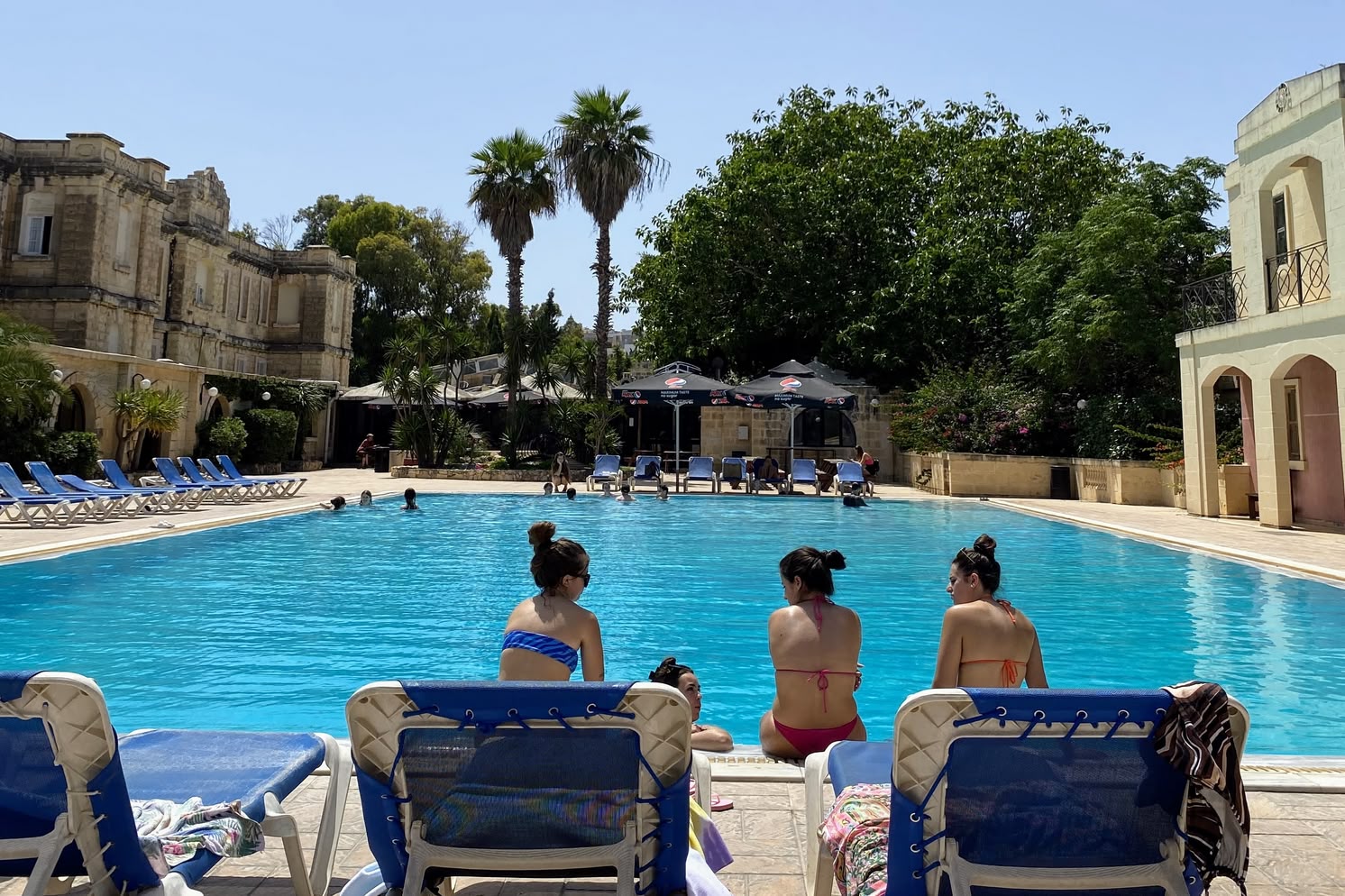 Three women sitting poolside on loungers at Sprachcaffe Malta, with historic buildings and palm trees in the background.