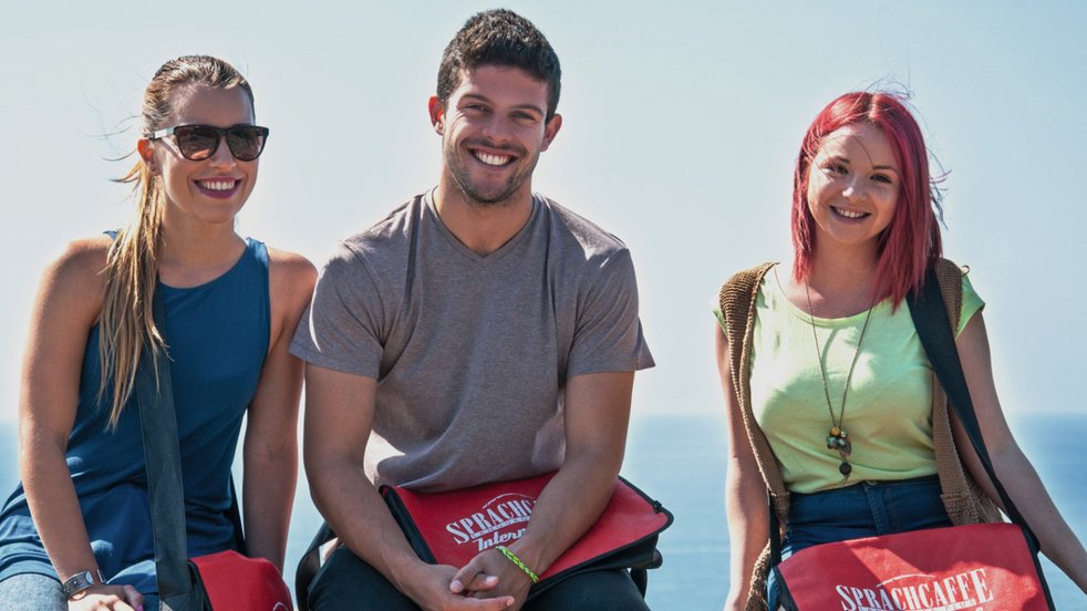 Three students with Sprachcaffe bags sitting on a railing, smiling with the sea in the background in Málaga, Spain.