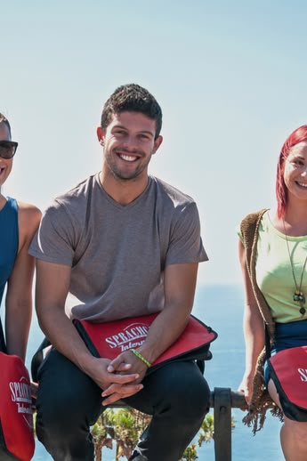 Three students with Sprachcaffe bags sitting on a railing, smiling with the sea in the background in Málaga, Spain.