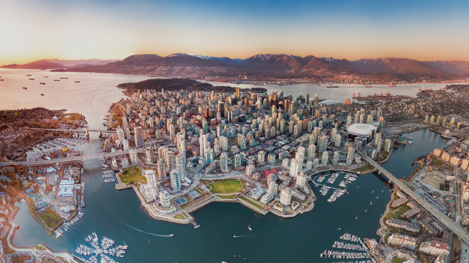 Vista aérea do centro de Vancouver, Canadá, com montanhas, portos e pontes ao redor, ao pôr do sol.