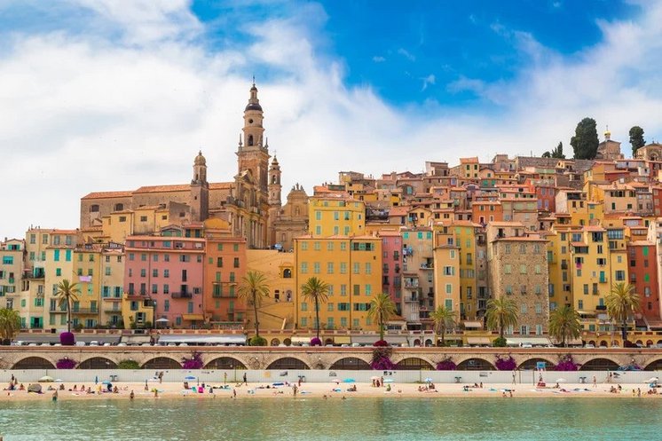 **ALT:** Kolorowe budynki i wieża kościoła w Menton, Francja, z widokiem na plażę i turkusowe wody Morza Śródziemnego, pod błękitnym niebem.