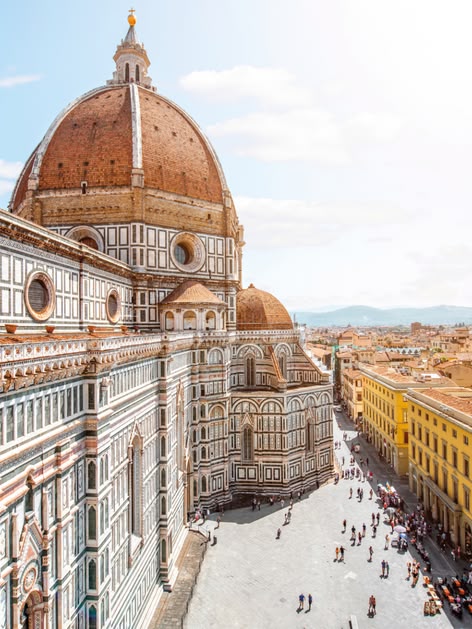 Zdjęcie przedstawia słynną katedrę Santa Maria del Fiore we Florencji, we Włoszech. Znana również jako Duomo di Firenze, katedra ta jest jednym z najbardziej rozpoznawalnych zabytków architektury renesansowej na świecie. Zbudowana w XIV i XV wieku, jej charakterystyczną cechą jest ogromna kopuła zaprojektowana przez Filippo Brunelleschiego. Na pierwszym planie widoczna jest fasada katedry, wykonana z marmuru w różnych kolorach, a w tle rozciąga się widok na urokliwe uliczki Florencji i charakterystyczne dachy budynków tego historycznego miasta.