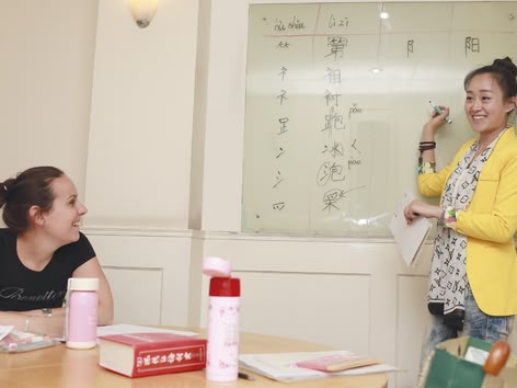 Teacher explaining Chinese characters on a whiteboard while a student listens and smiles.