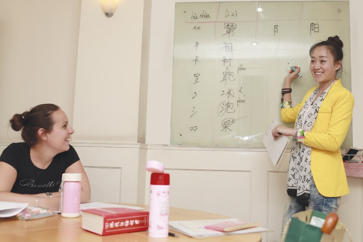 Teacher explaining Chinese characters on a whiteboard while a student listens and smiles.