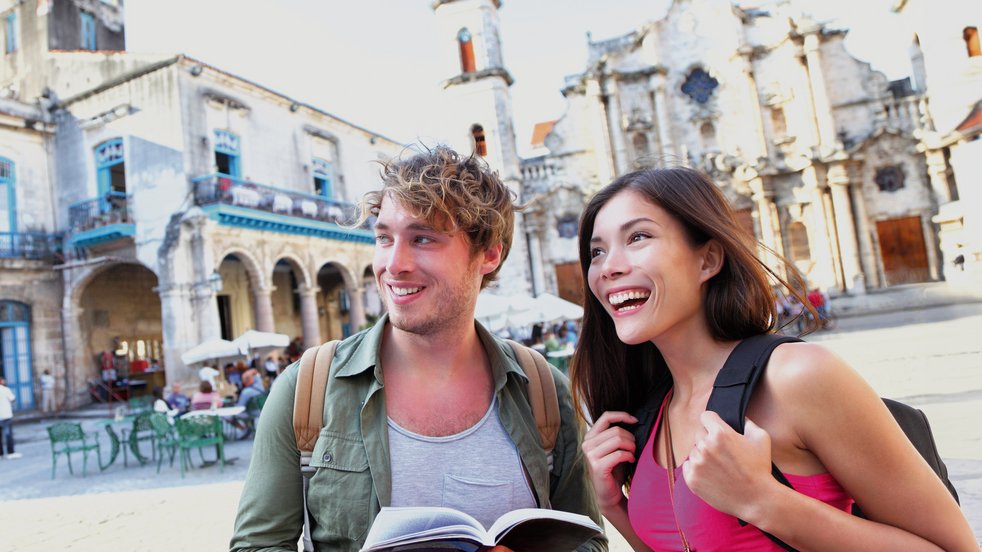 Two tourists exploring Old Havana, Cuba, holding a guidebook and smiling in a historic square with colonial architecture.