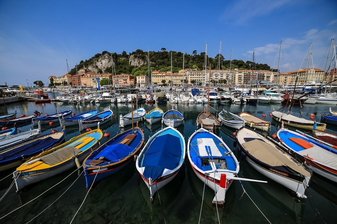 Colourful boats docked in the Port of Nice, France, with hillside buildings and a marina in the background.
