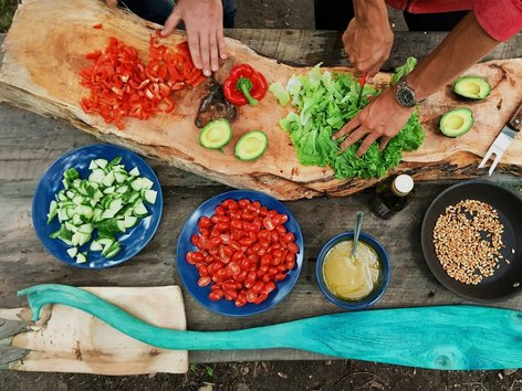 An overhead view (flat lay) on a rustic wooden table showing hands chopping lettuce and red peppers, with blue bowls containing diced cucumbers and cherry tomatoes, roasted nuts, dressing, avocados, and a large turquoise wooden spoon.