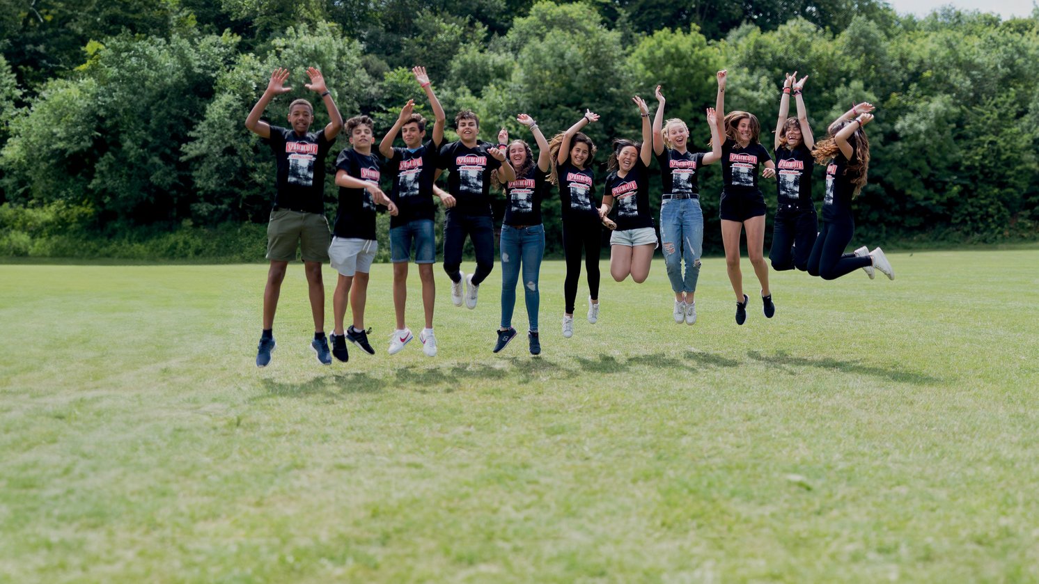 Sprachcaffe students in England jumping mid-air on a green field, wearing matching shirts and enjoying the outdoors.