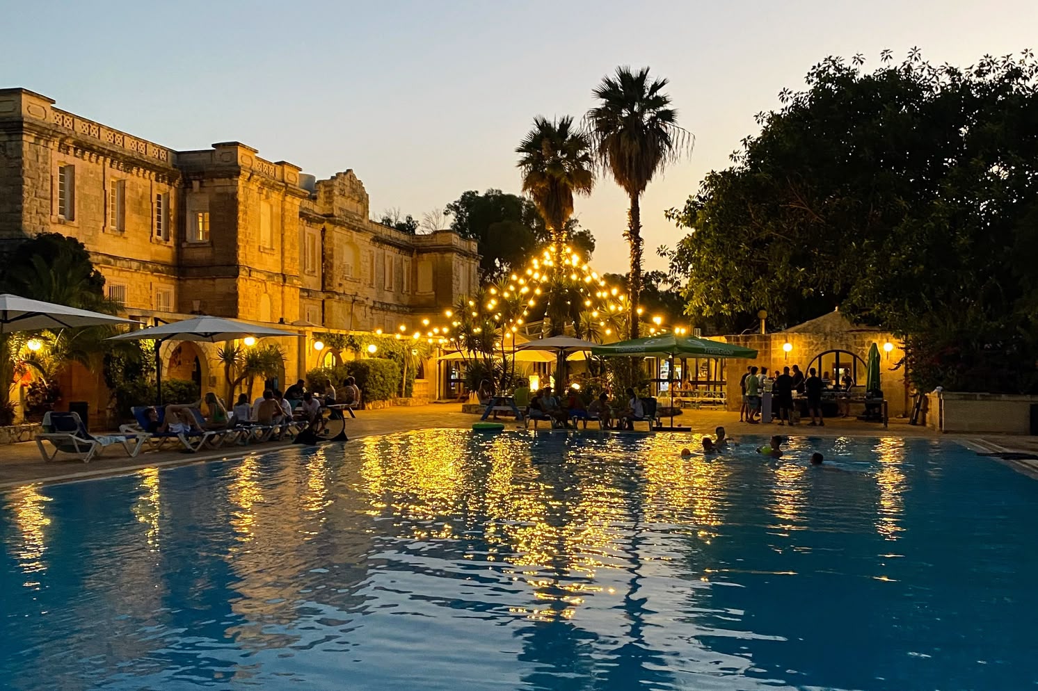 Evening view of the Sprachcaffe Malta pool area lit by string lights, with students gathered around and historic buildings in the background.