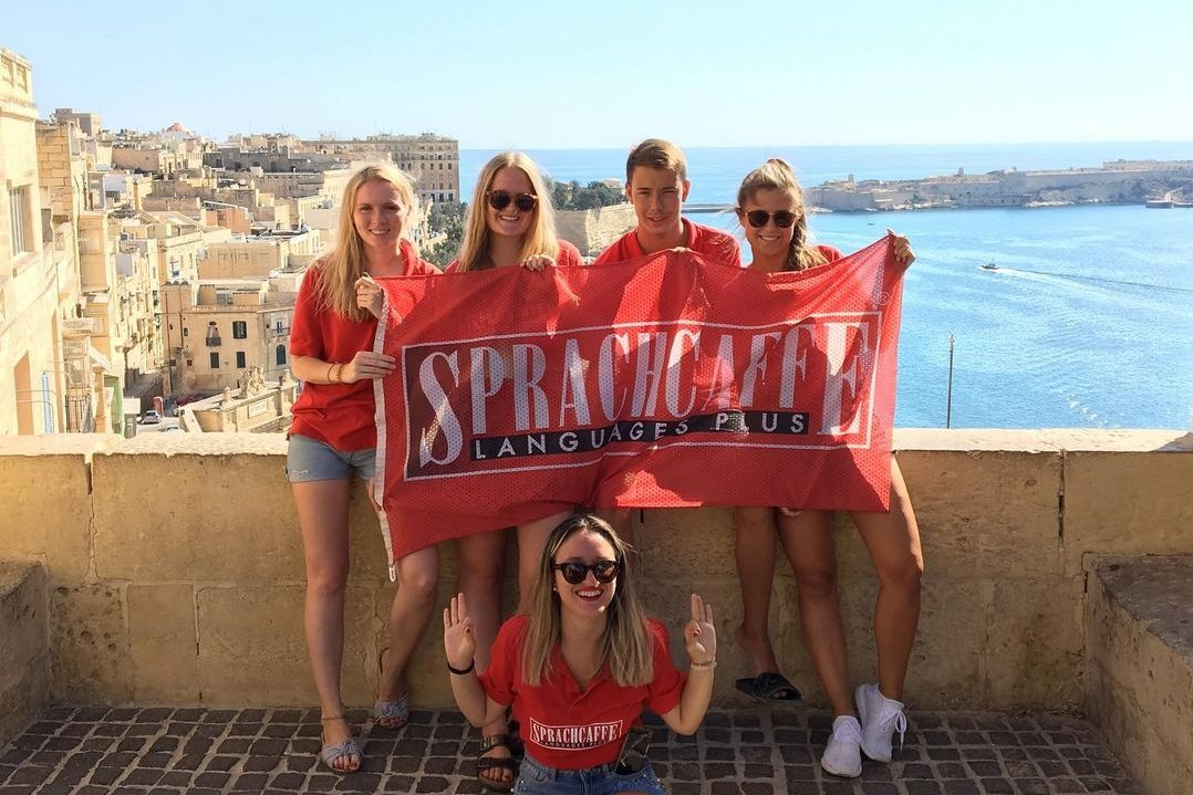 Grupa studentów Sprachcaffe trzymających czerwoną flagę szkoły na tle panoramy Valletty i morza.