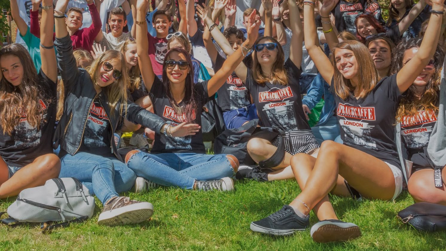 Grupo de alunos da Sprachcaffe sentados na grama em Londres, sorrindo e levantando os braços durante uma atividade ao ar livre.