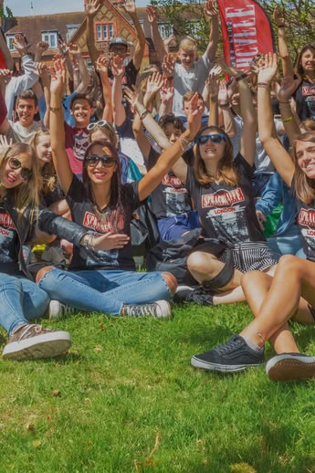 Group of Sprachcaffe students sitting on the grass in London, smiling and raising their arms during an outdoor activity.