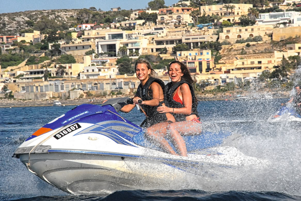 Two young women smiling while riding a jet ski along Malta's coast.