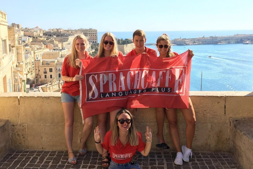 Grupa studentów Sprachcaffe trzymających czerwoną flagę szkoły na tle panoramy Valletty i morza.