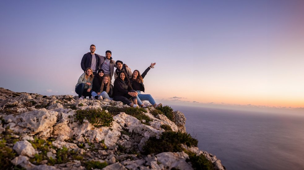 Group of young adults on a rocky cliff in Malta at sunset, overlooking the sea and enjoying the view.
