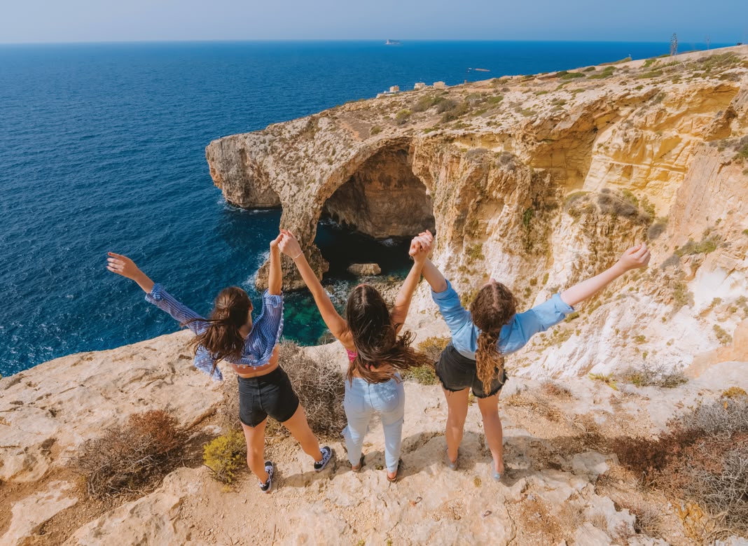 Three girls raising arms on cliffs overlooking the Blue Grotto.